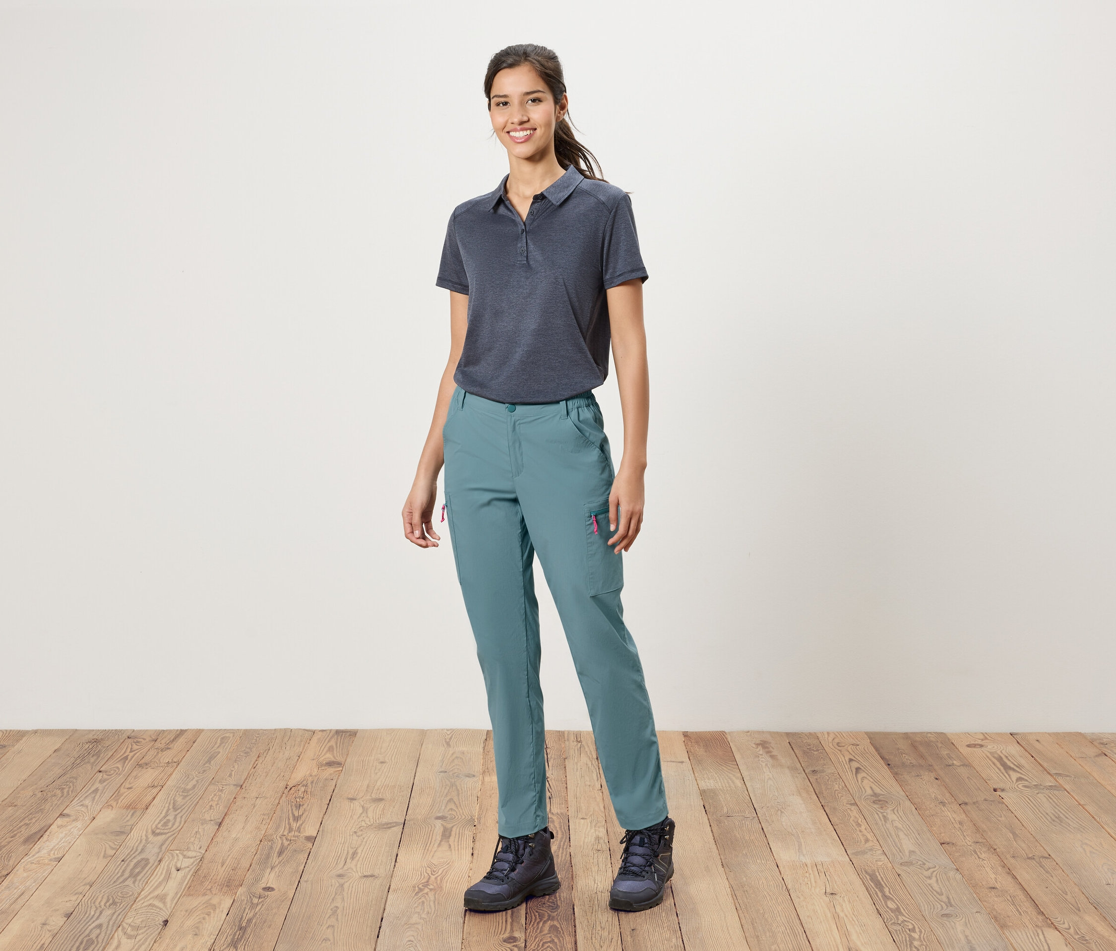 Žena stojí před bílou zdí a má na sobě modré polokošile, modré kalhoty a trekové boty.