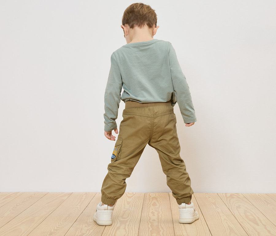 Dítě stojí zády ke kameře, má na sobě zelené tričko s dlouhým rukávem, khaki kalhoty a bílé tenisky.