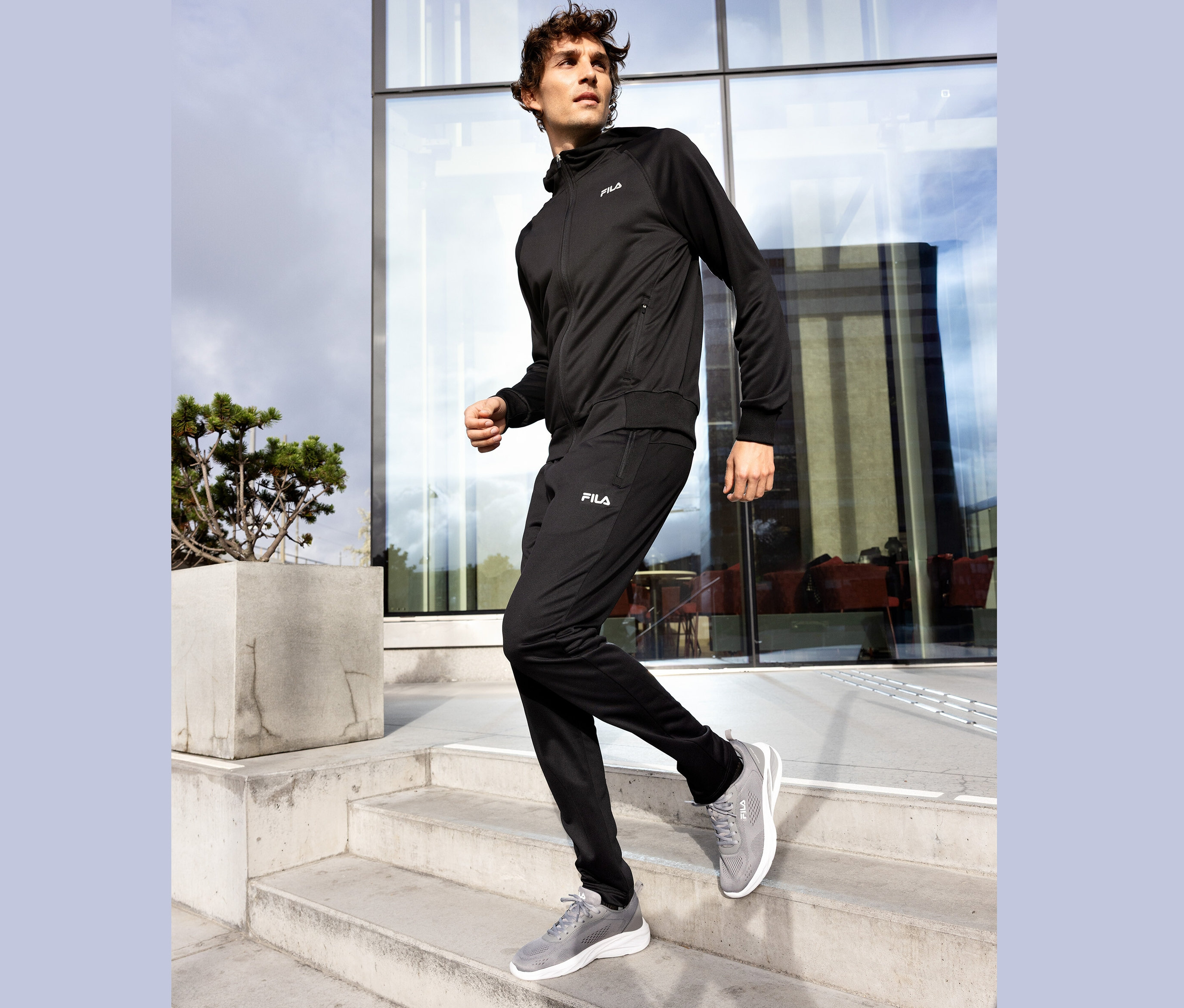 Muž v černé teplákové soupravě Fila a šedých teniskách schází po betonových schodech.