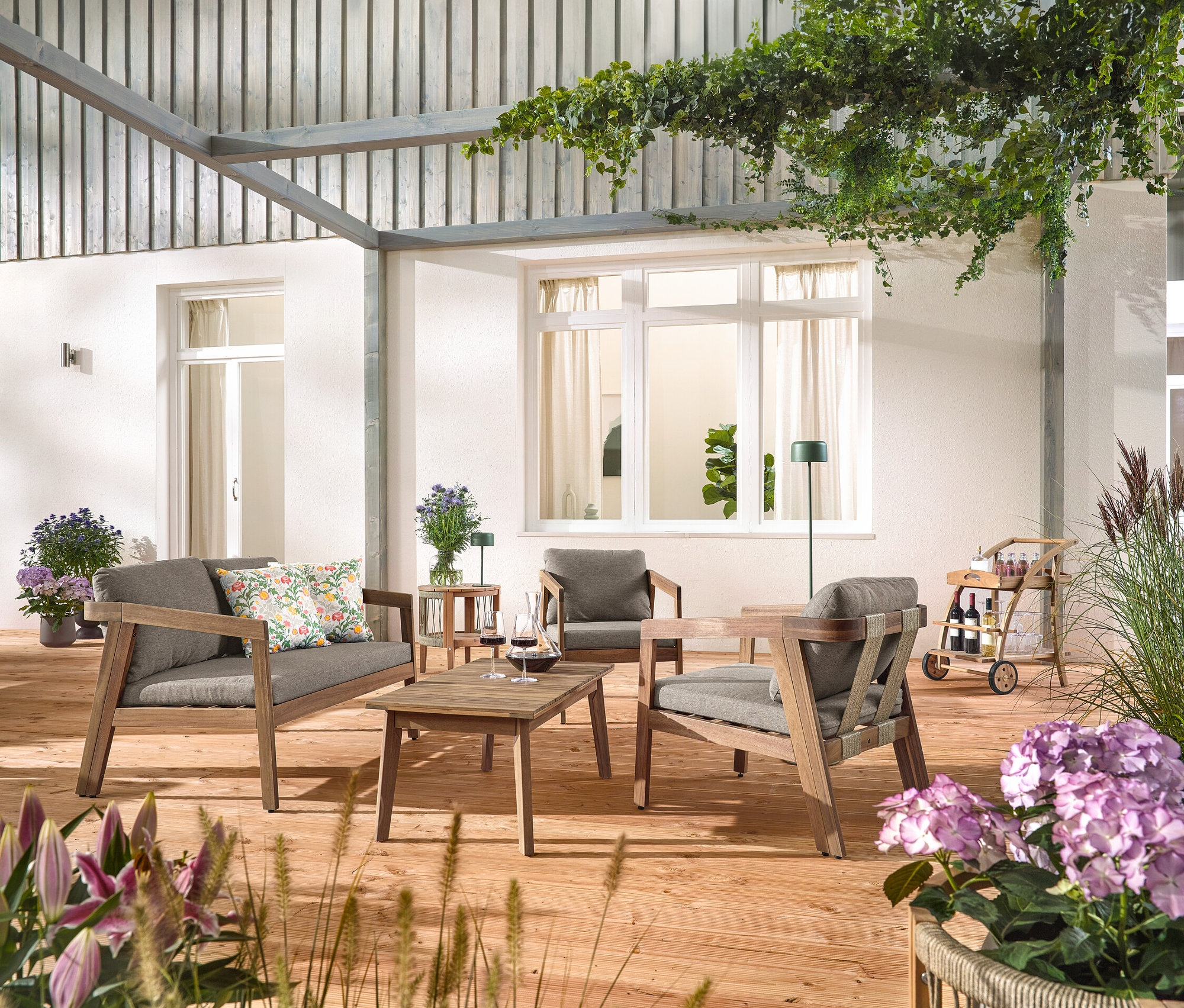 Na dřevěné terase stojí 4dílná zahradní sedací souprava s textilním výpletem, se 2 venkovními polštáři s květinovým vzorem, barový vozík s odnímatelným podnosem a zahradní odkládací stolek s odnímatelnou deskou, průměr cca 50 cm. Vidět je také solární dobíjecí stolní lampa, džunglová zelená a solární dobíjecí stojací lampa, džunglová zelená.