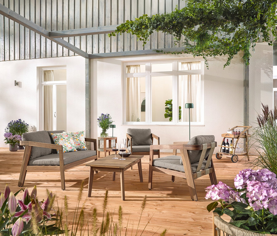 Na dřevěné terase stojí 4dílná zahradní sedací souprava s textilním výpletem, se 2 venkovními polštáři s květinovým vzorem, barový vozík s odnímatelným podnosem a zahradní odkládací stolek s odnímatelnou deskou, průměr cca 50 cm. Vidět je také solární dobíjecí stolní lampa, džunglová zelená a solární dobíjecí stojací lampa, džunglová zelená.