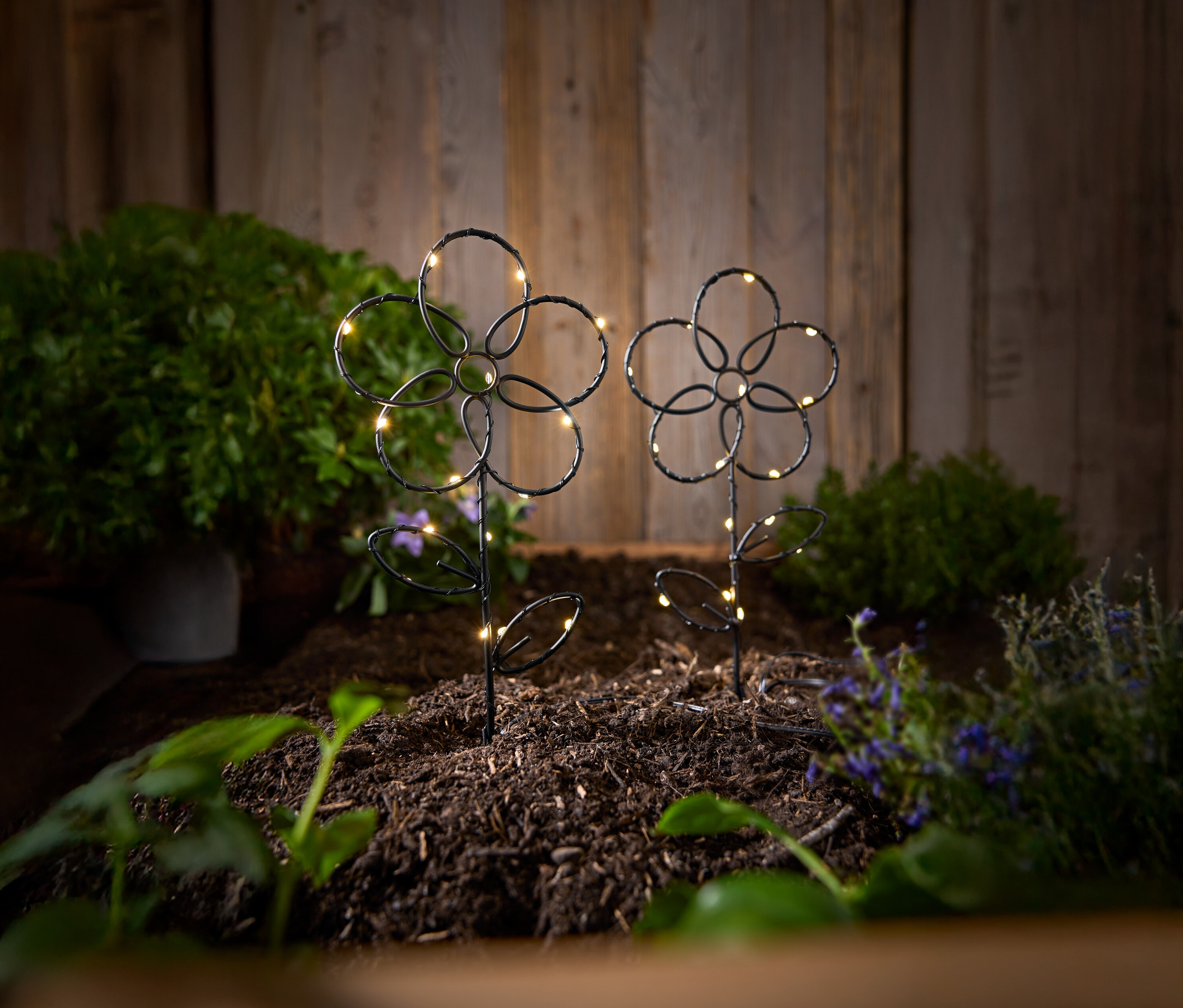 Dva černé, květinové zahradní kolíky s LED světly stojí v zemi před dřevěnou stěnou.