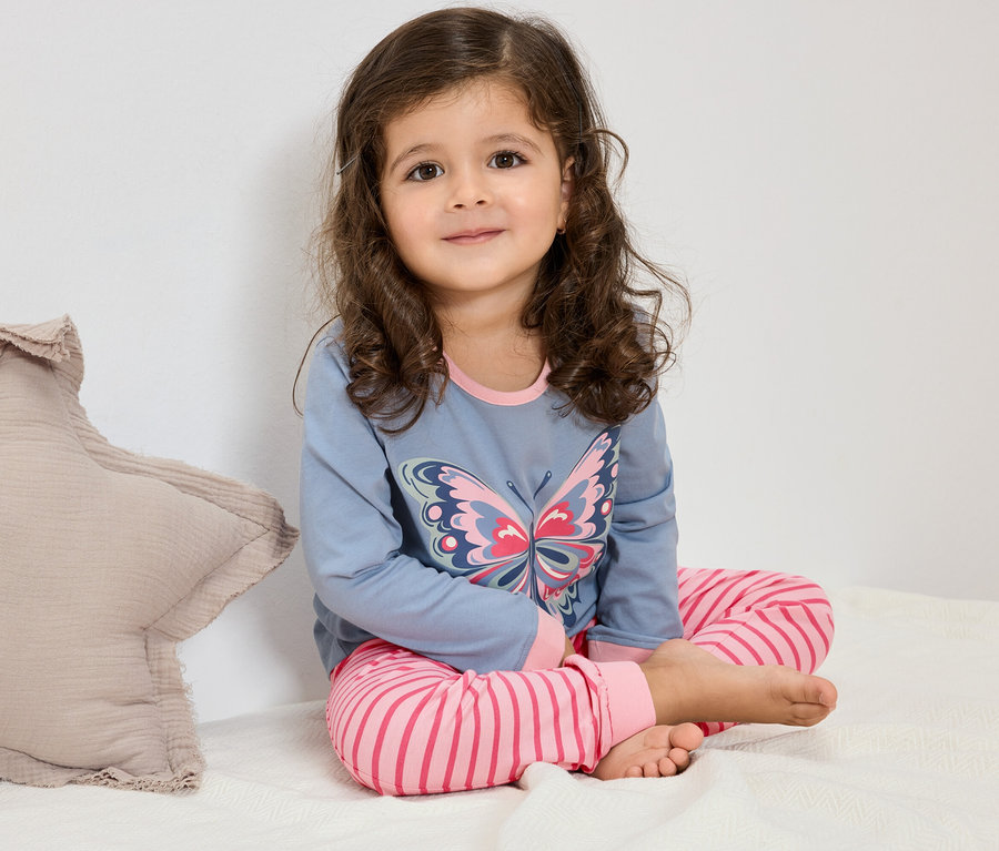 Dívka s kudrnatými vlasy sedí na posteli a má na sobě modré pyžamo s motýlem a růžové pruhované kalhoty.