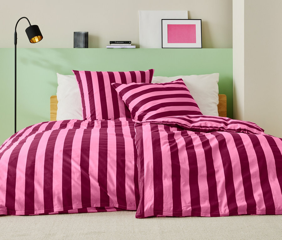 Postel s růžovo-vínově červeným pruhovaným ložním prádlem, dvěma polštáři a stojací lampou.
