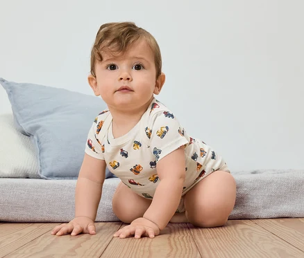 Kojence leze po dřevěné podlaze, oblečené v body s krátkým rukávem se vzorem stavebních vozidel.