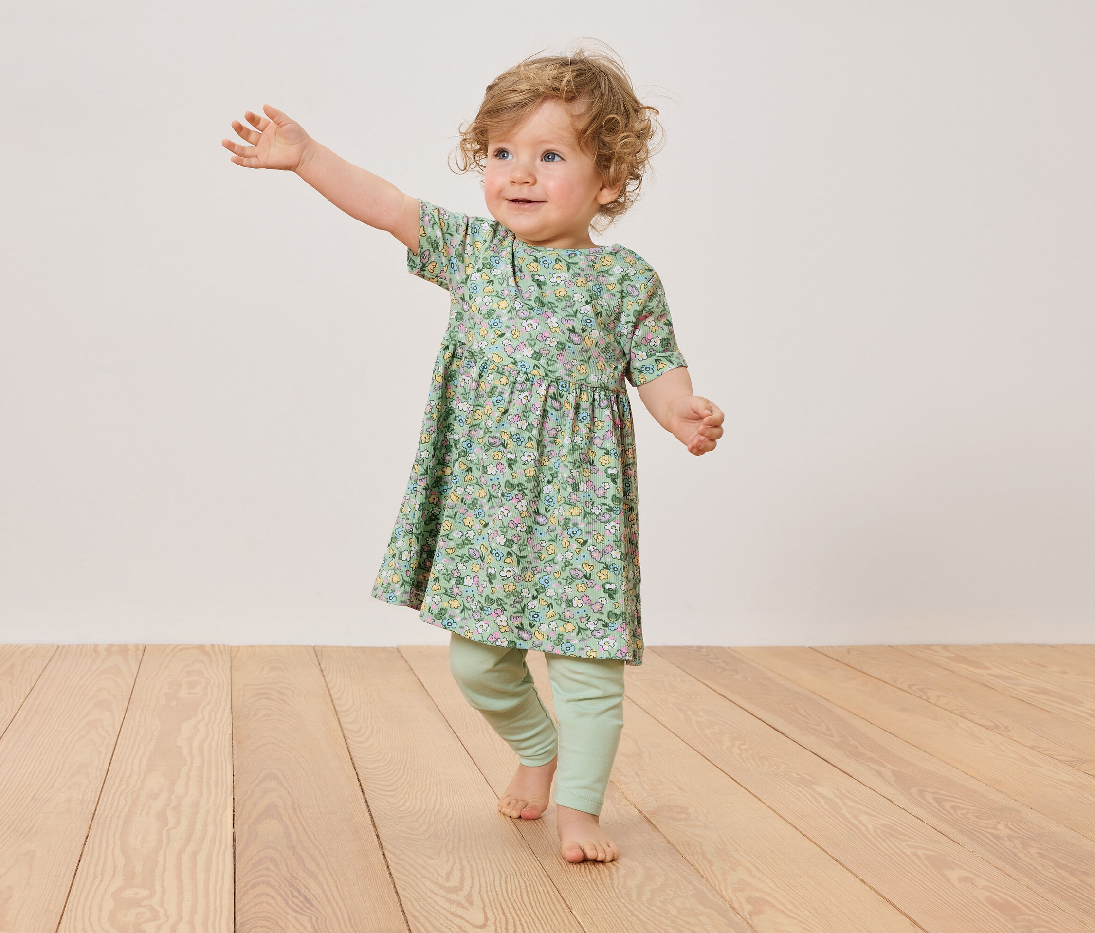 Batole s blond kudrlinkami má na sobě květované šaty a zelené legíny, stojí na dřevěné podlaze.