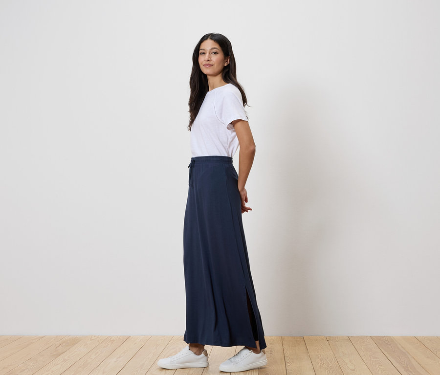 Žena stojí bokem, má na sobě bílé tričko, dlouhou tmavě modrou sukni a bílé tenisky.