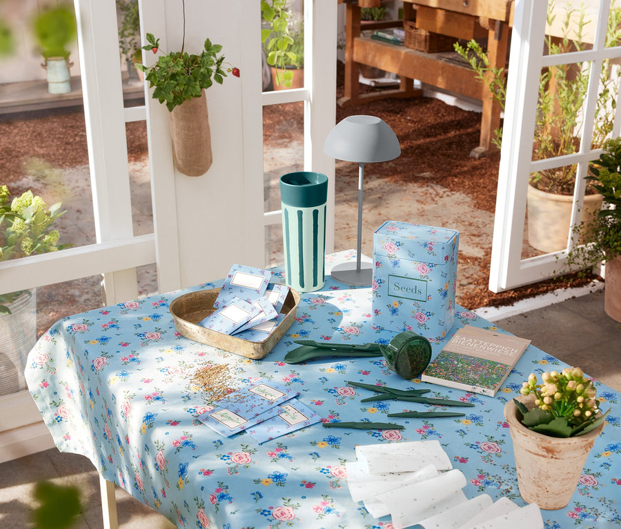 Šedá LED akumulátorová stolní lampa stojí na stole s květinovým ubrusem, vedle zahradního nářadí a semen.