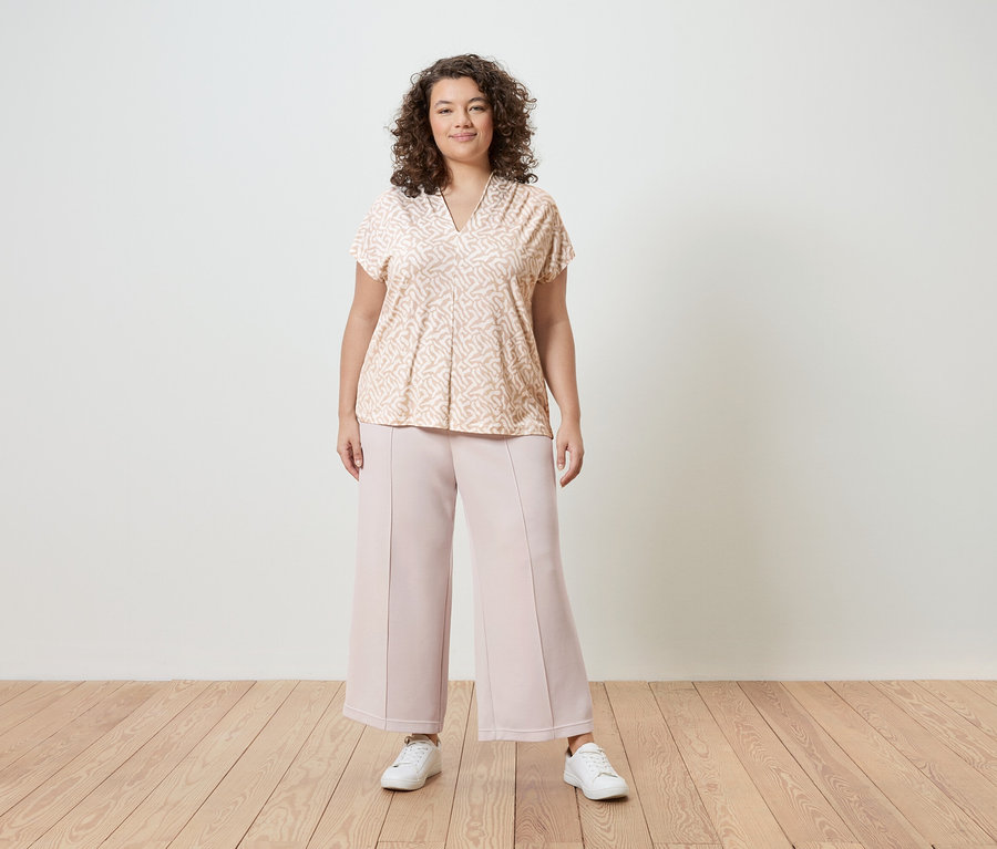 Žena s kudrnatými vlasy má na sobě světle růžovou vzorovanou halenku, růžové kalhoty culottes a bílé tenisky před bílou stěnou.