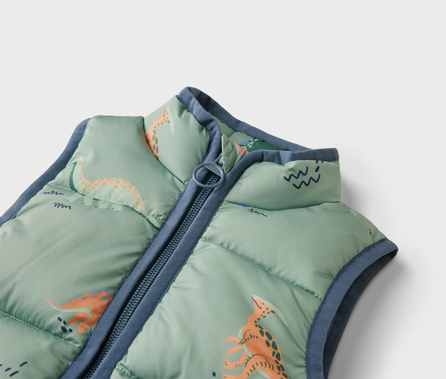 Detail prošívané světle zelené vesty s motivem dinosaurů a modrým zipem.