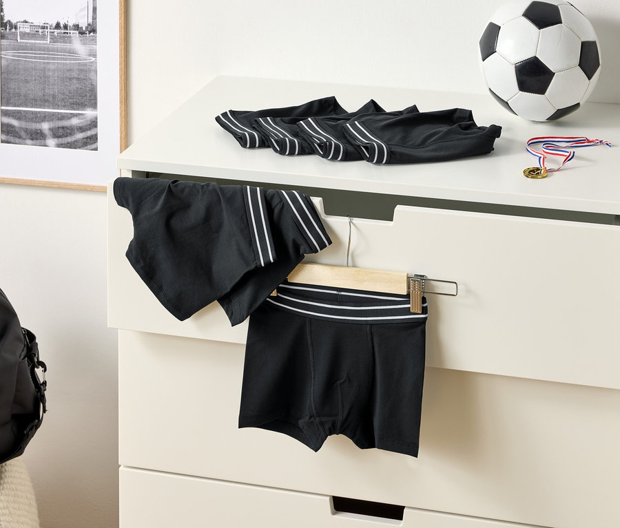 Na komodě leží černé boxerky s bílými pruhy, fotbalový míč a medaile. Na ramínku visí sportovní šortky.