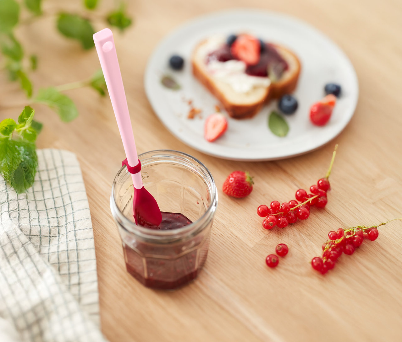 Sklenice džemu s růžovou lžičkou, toast s jahodami a borůvkami a rybíz na dřevěném stole.
