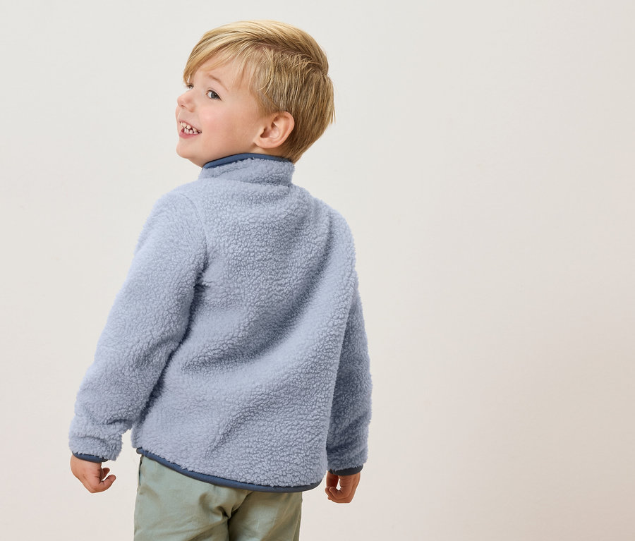 Malý chlapec s blond vlasy má na sobě světle modrou fleecovou bundu a světle zelené kalhoty, dívá se stranou.