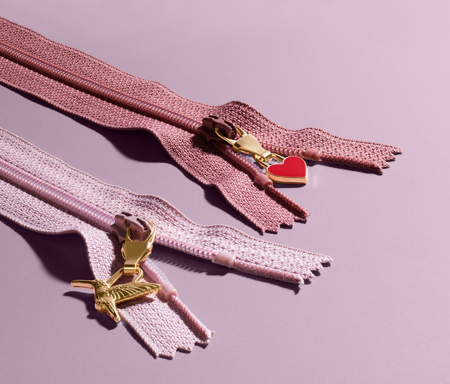 Dva zipy s ozdobnými jezdci na fialovém pozadí. Horní zip má červený přívěsek ve tvaru srdce, dolní zip má přívěsek ve tvaru kolibříka. Přívěsky na zip, 6 ks.