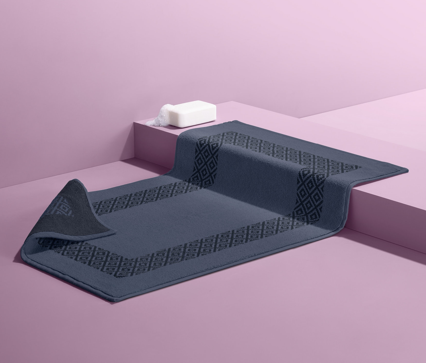 Modrá oboustranná koupelnová předložka s geometrickým vzorem leží na růžovém pozadí. Na ní je bílé mýdlo.