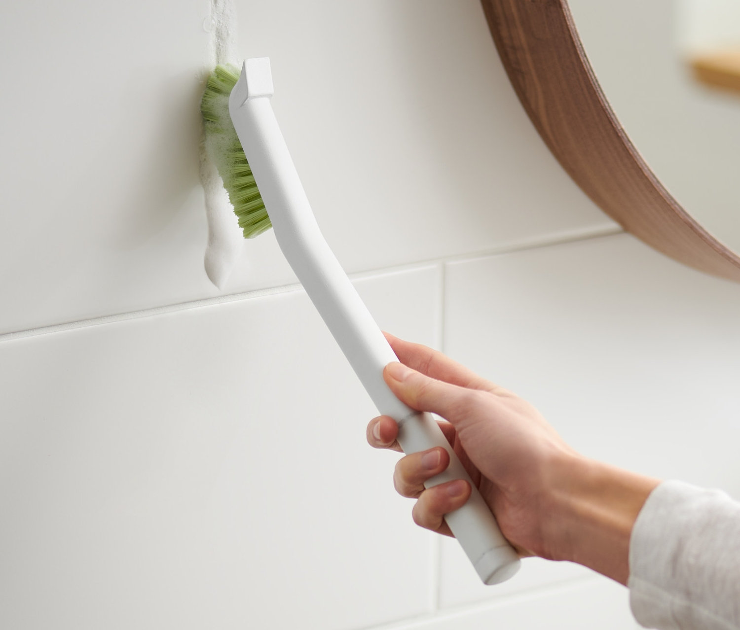 Žena čistí spáry mezi bílými dlaždicemi dlouhým kartáčem na spáry.