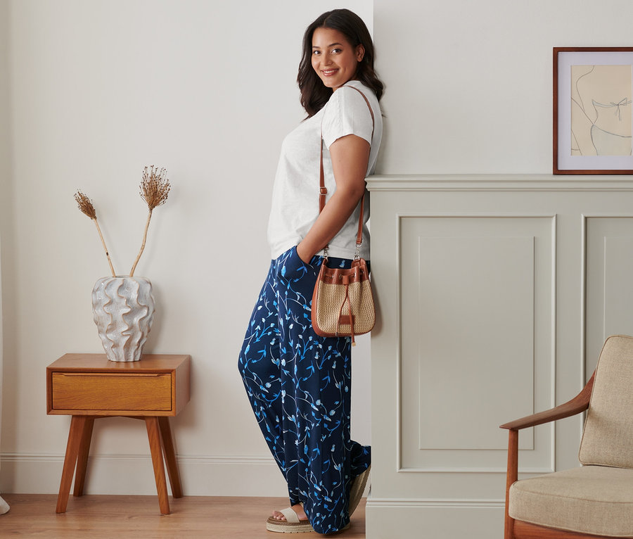 Žena opřená o zeď pózuje v širokých kalhotách, bílém tričku a kožených sandálech na platformě. Přes rameno má přehozenou kabelku.