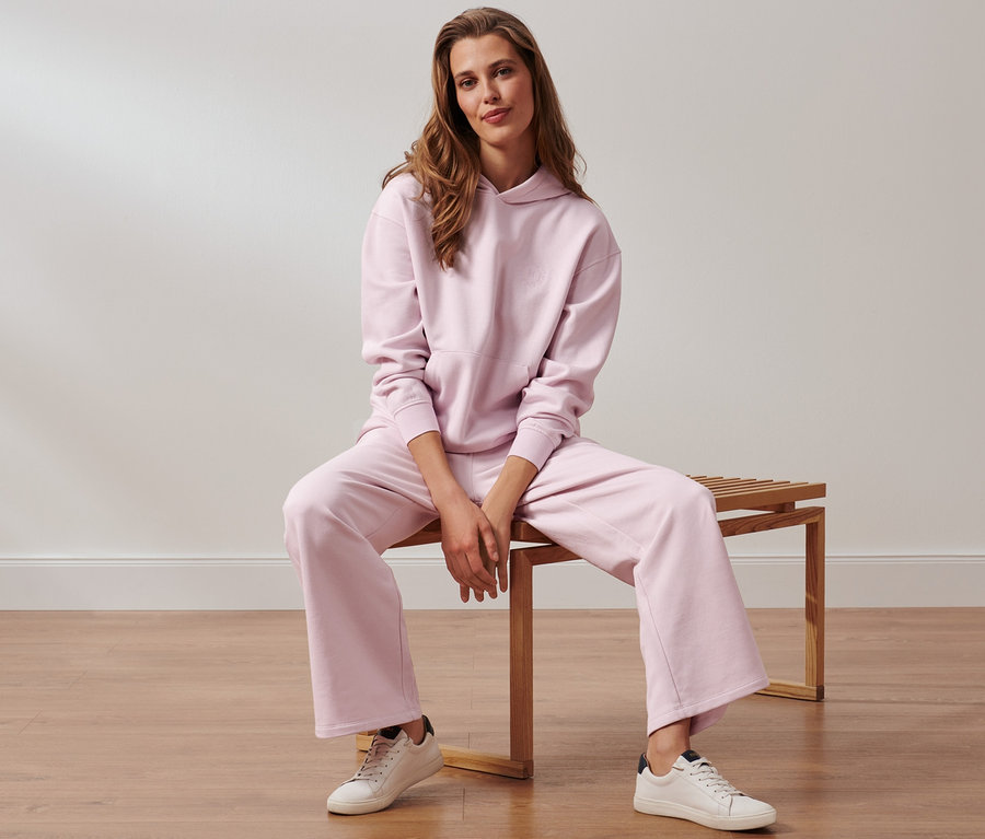 Žena sedí na lavici v růžových teplákách a mikině s kapucí.