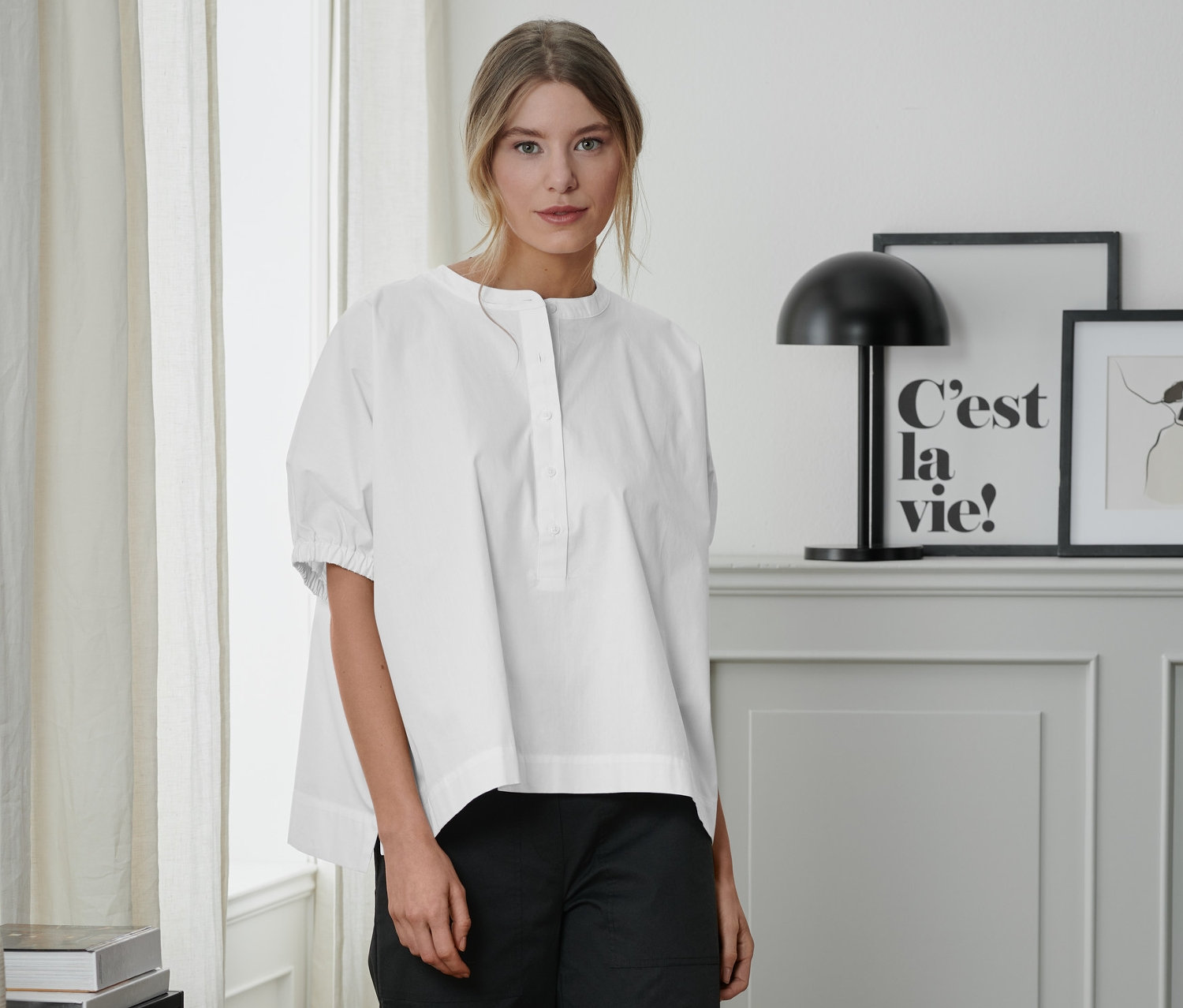 Žena stojí a má na sobě bílou popelínovou halenku a černé kalhoty.