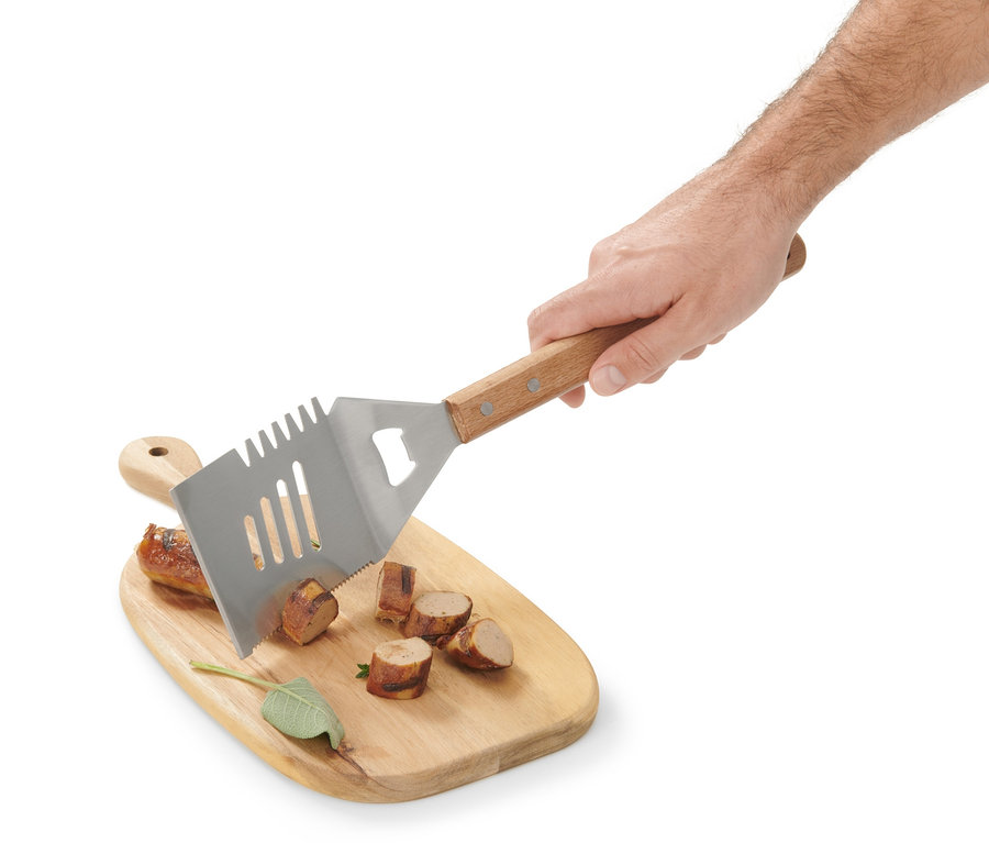 Ruka drží multifunkční BBQ náčiní nad dřevěným prkénkem s nakrájenou klobásou.