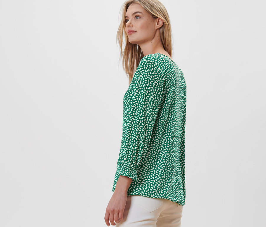 Blondýna pózuje bokem a má na sobě zelené halenkové triko s potiskem.