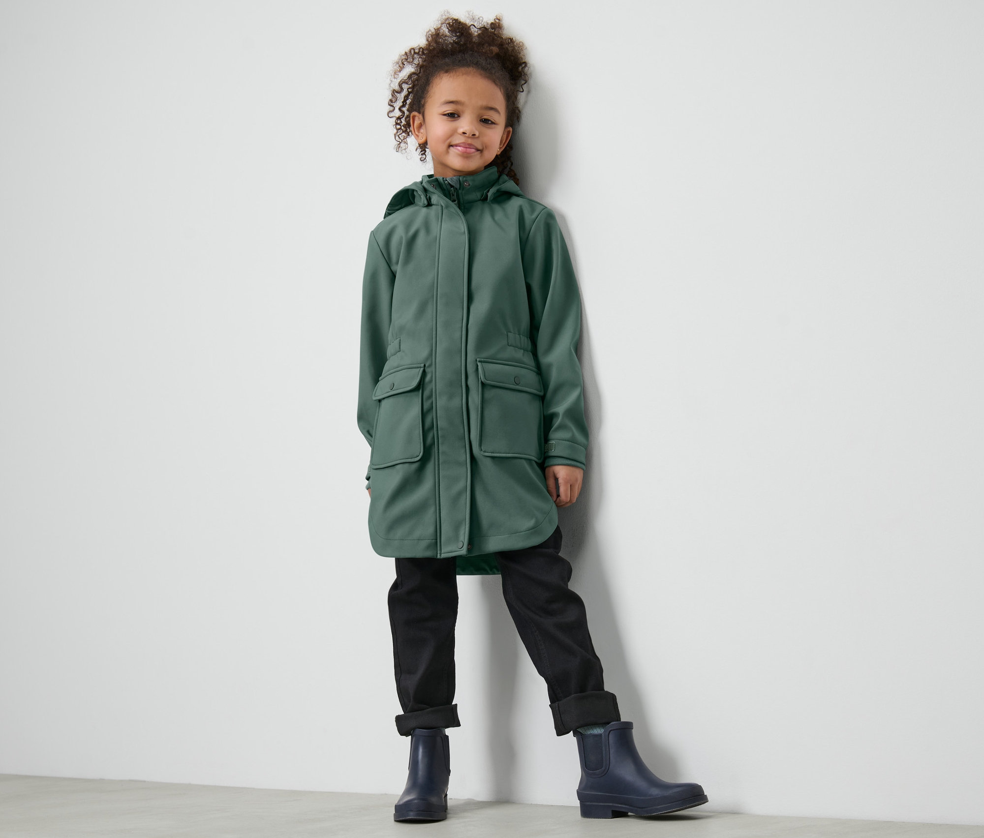 Dívka stojí opřená o zeď v dětském softshellovém kabátu, černých kalhotách a modrých botách.