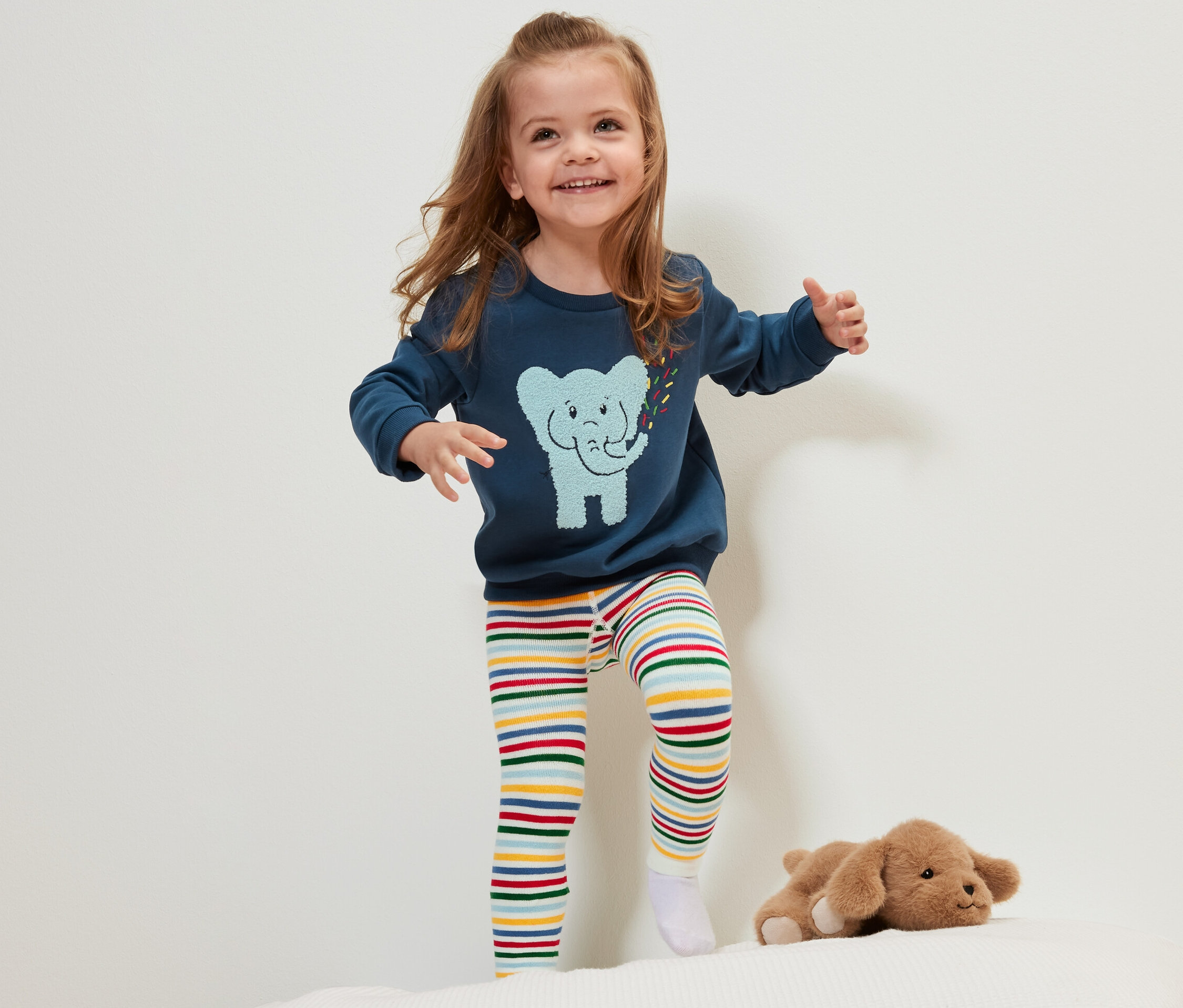 Usmívající se holčička stojí na posteli a má na sobě termolegíny Dětské termolegíny, 3 ks s barevnými pruhy.