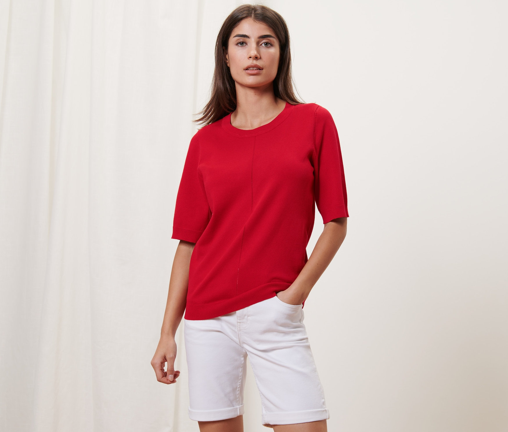 Žena pózuje v červeném svetříku z jemného úpletu a bílých šortkách.
