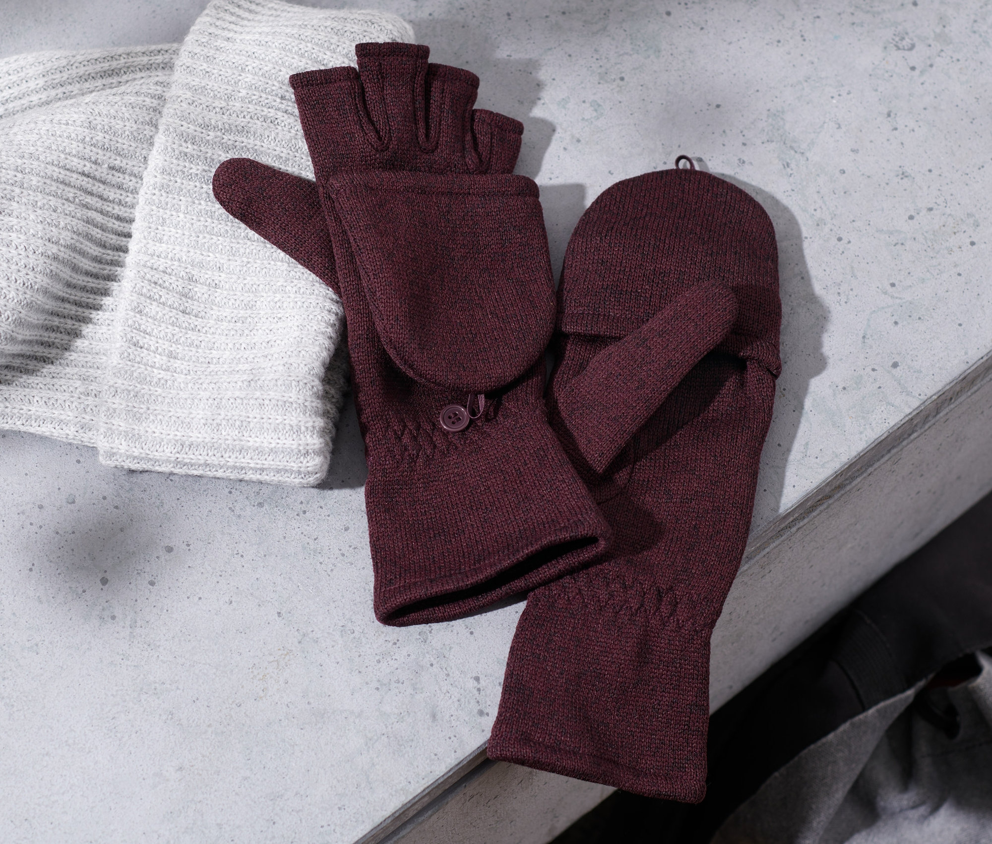 Pletená čepice leží vedle bordó odklápěcích palčáků z pleteného fleecu.