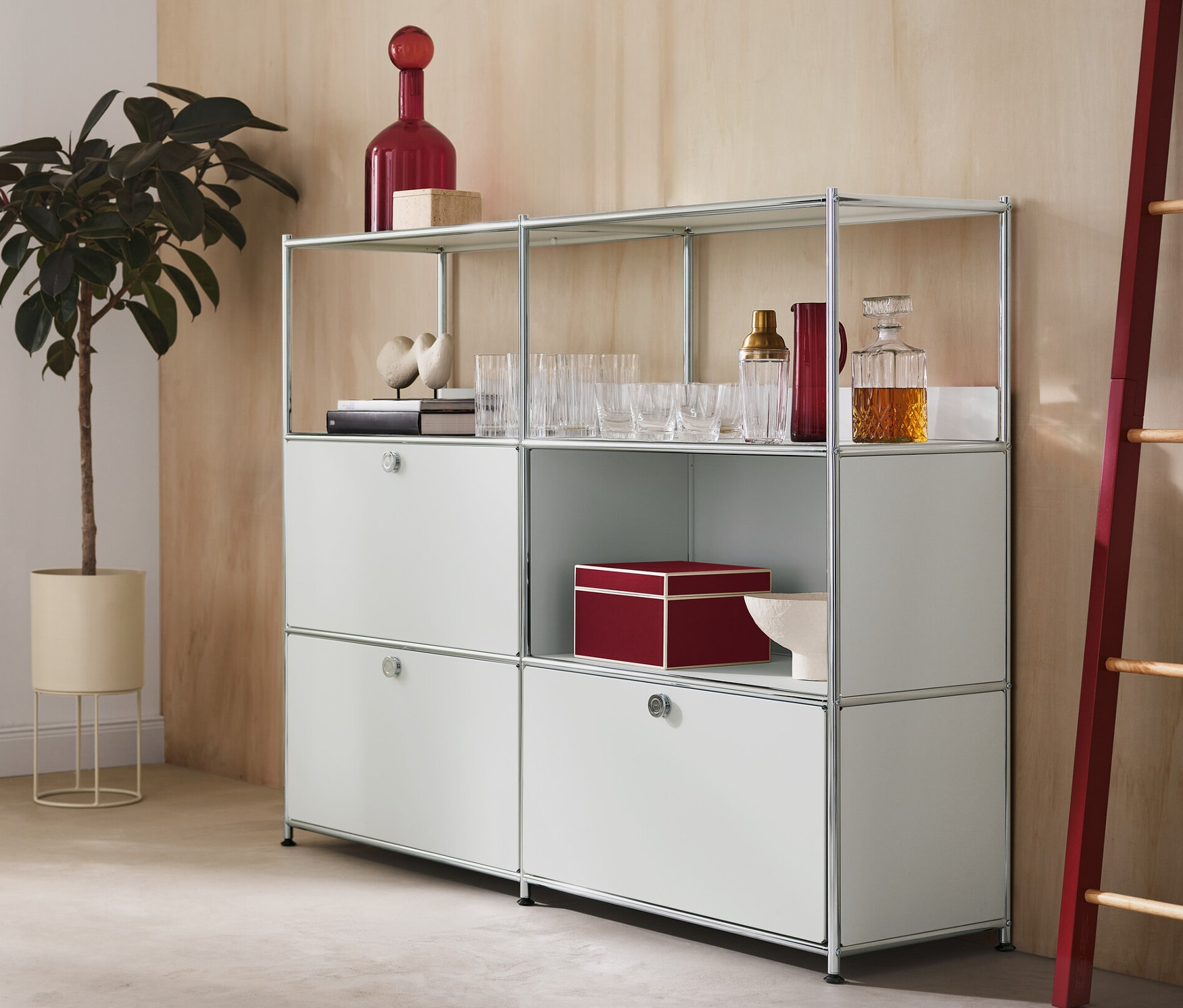 V místnosti je kulatý květináč s rostlinou, kovová komoda »CN3« se 2 výklopnými dvířky a 1 zásuvkou a dekorativní žebřík s červenými bočními prvky.