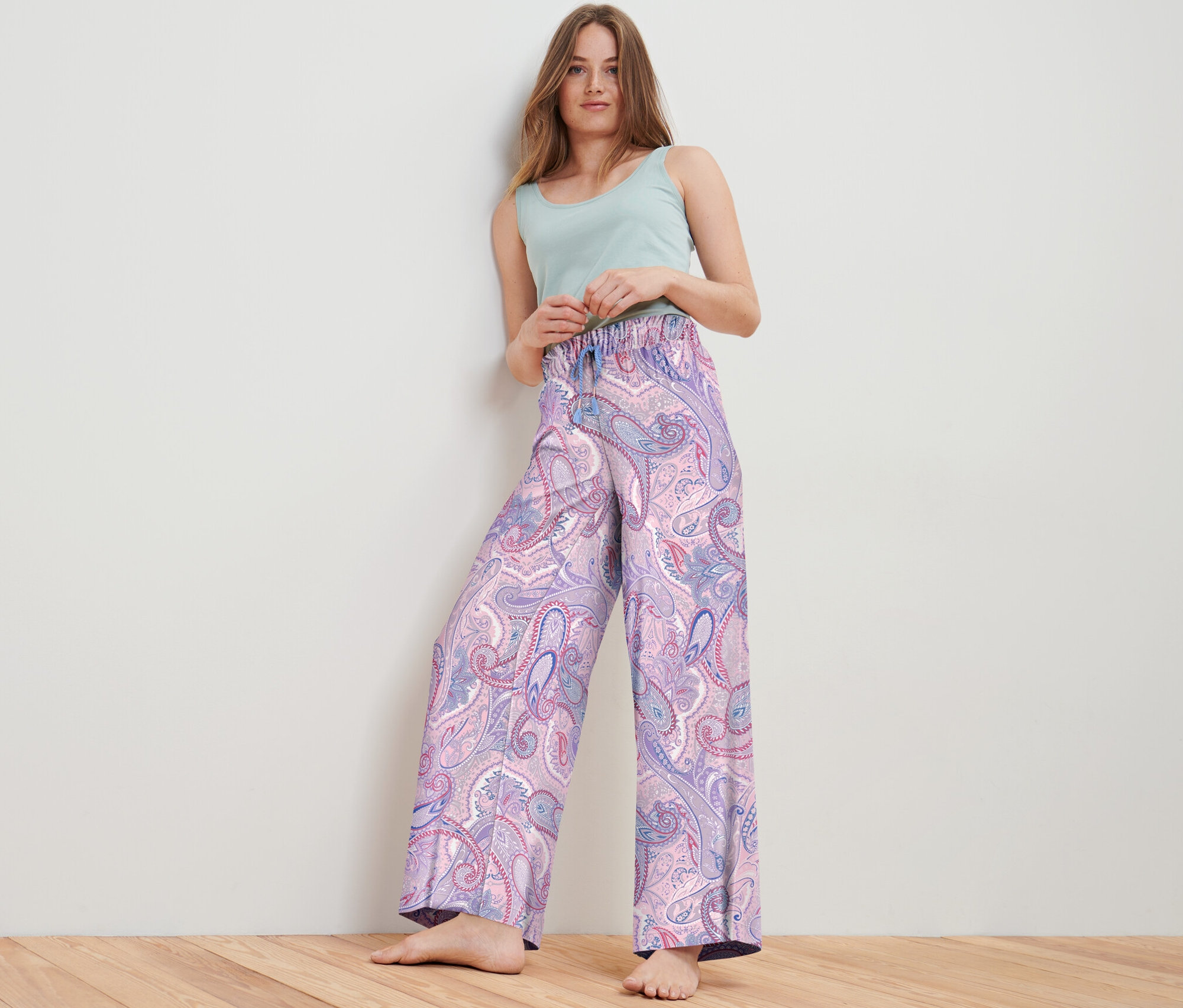 Žena pózuje ve studiu a má na sobě tkané kalhoty s paisley vzorem a světle zelený top.