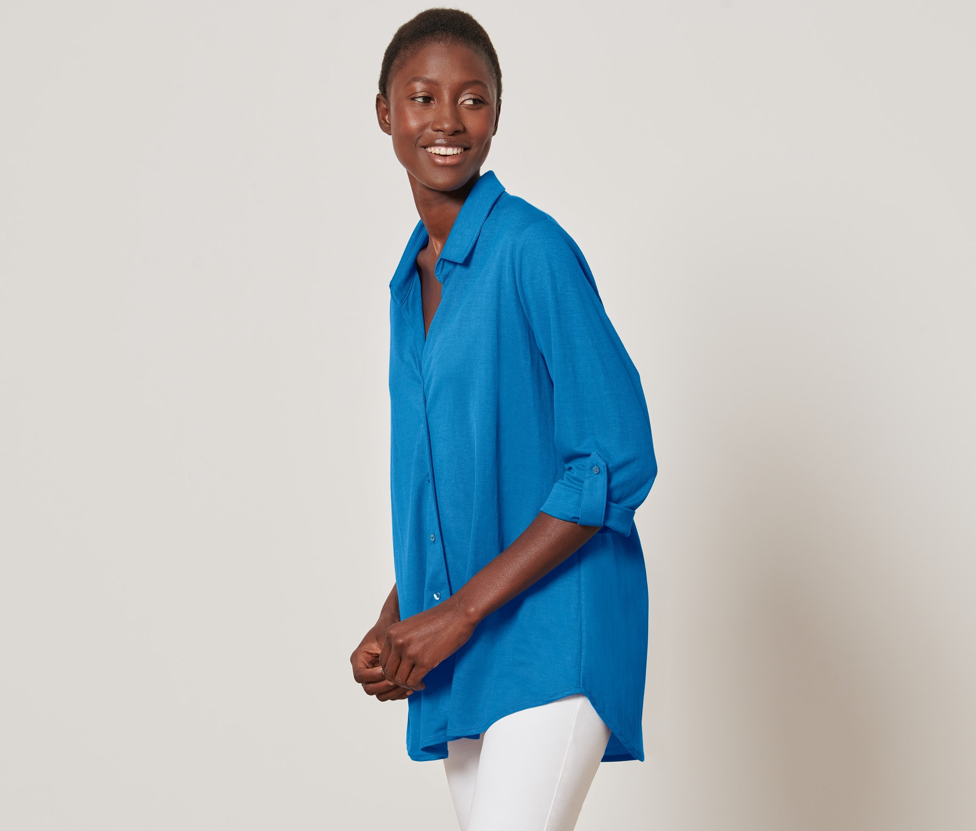 Usmívající se žena pózuje v azurově modré žerzejové halence piké a bílých kalhotách.