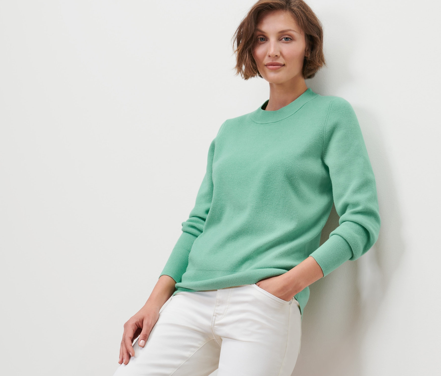 Žena opřená o zeď má na sobě pletený svetr a bílé džíny.