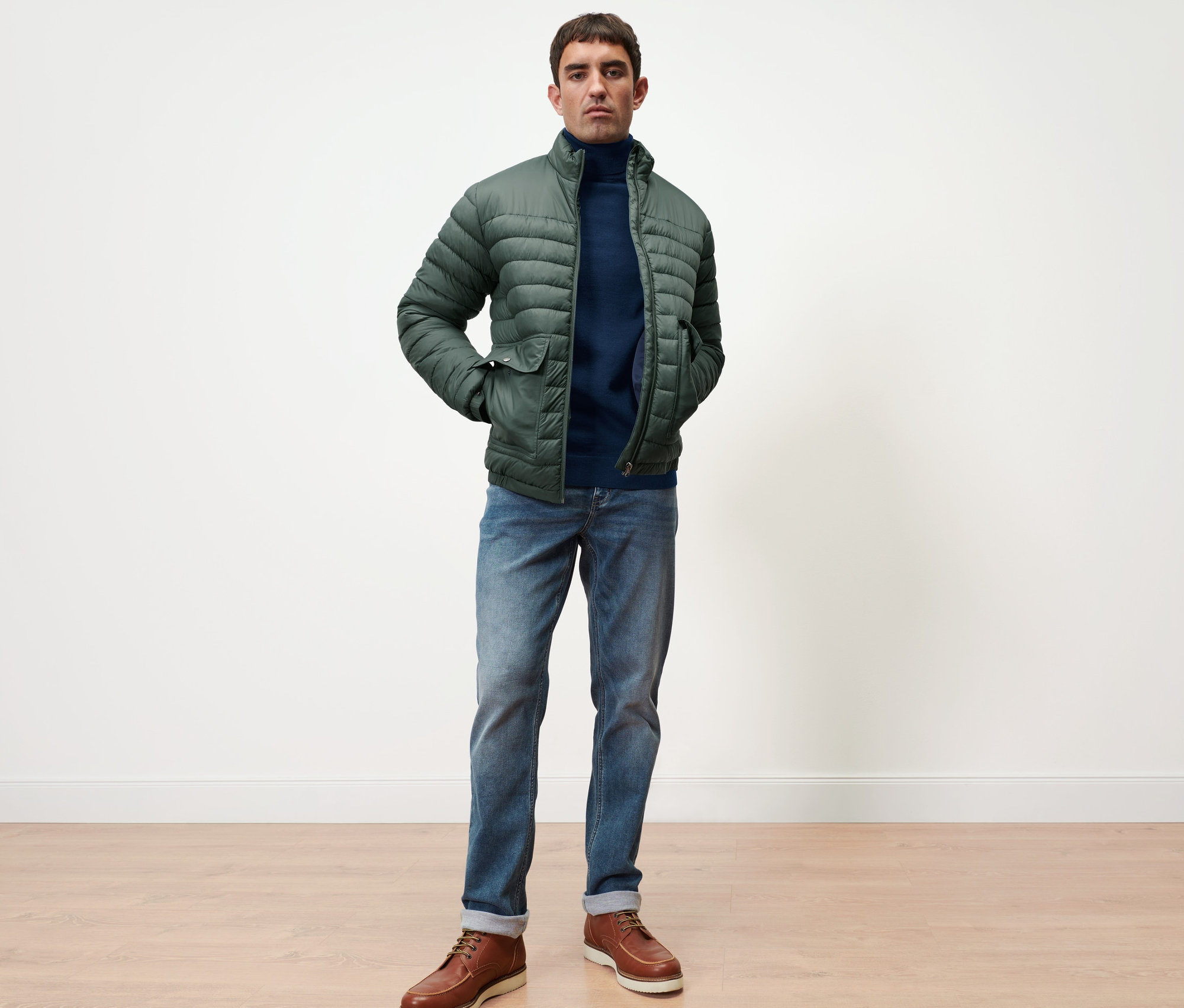 Muž stojí a má na sobě námořnicky modrý svetr z merino vlny a zelenou prošívanou bundu.