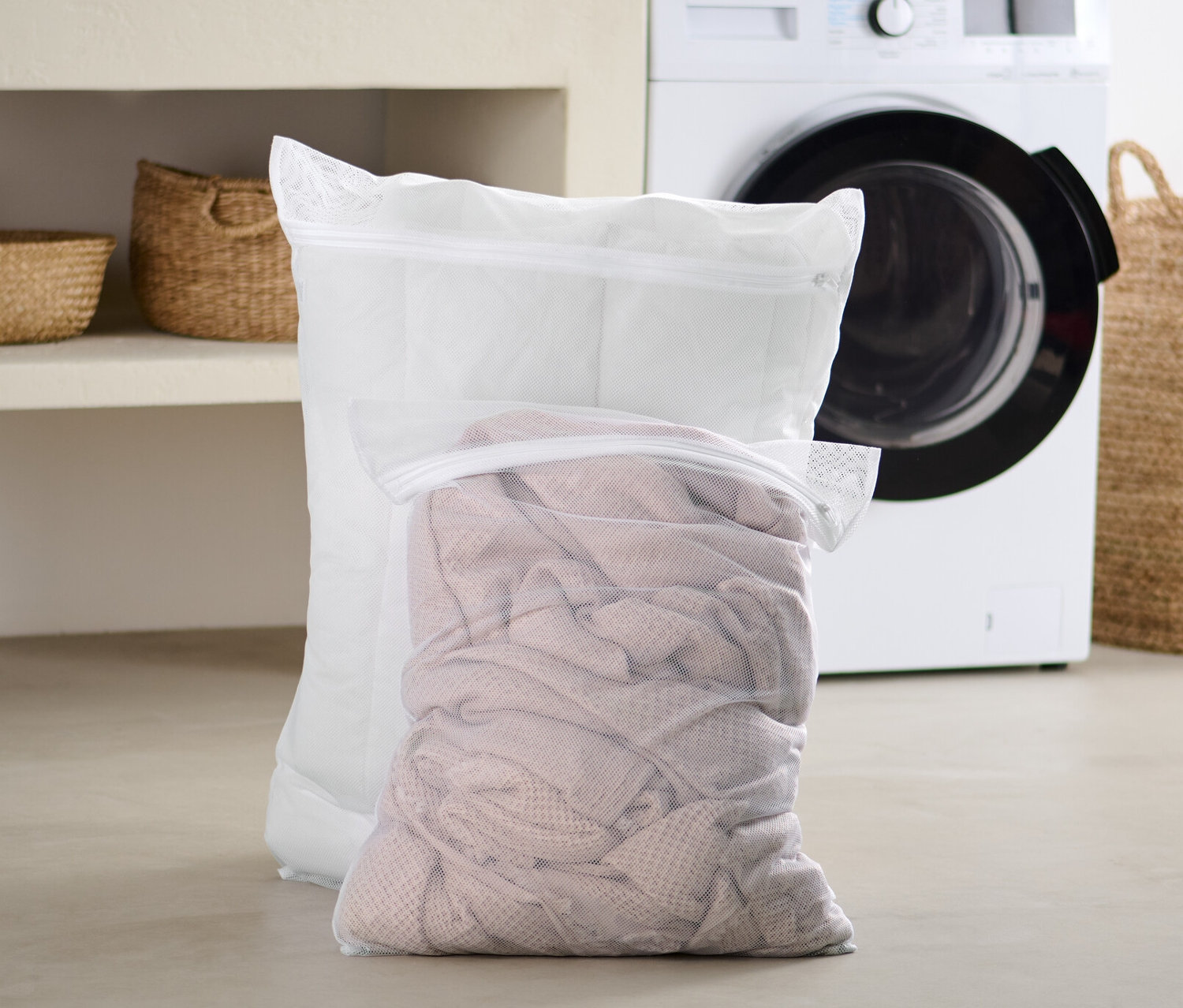 Dvě extra velké síťky na praní prádla stojí na podlaze v koupelně před pračkou.