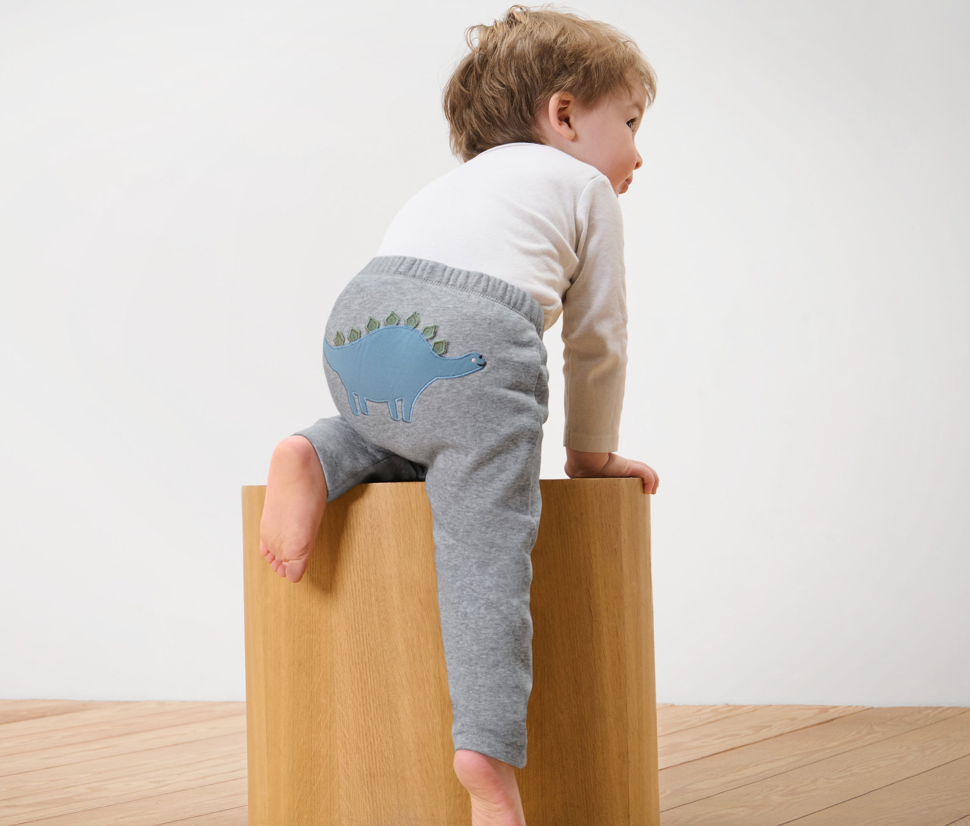 Dítě sedí na dřevěné kostce a má na sobě šedé tepláky s dinosaurem.