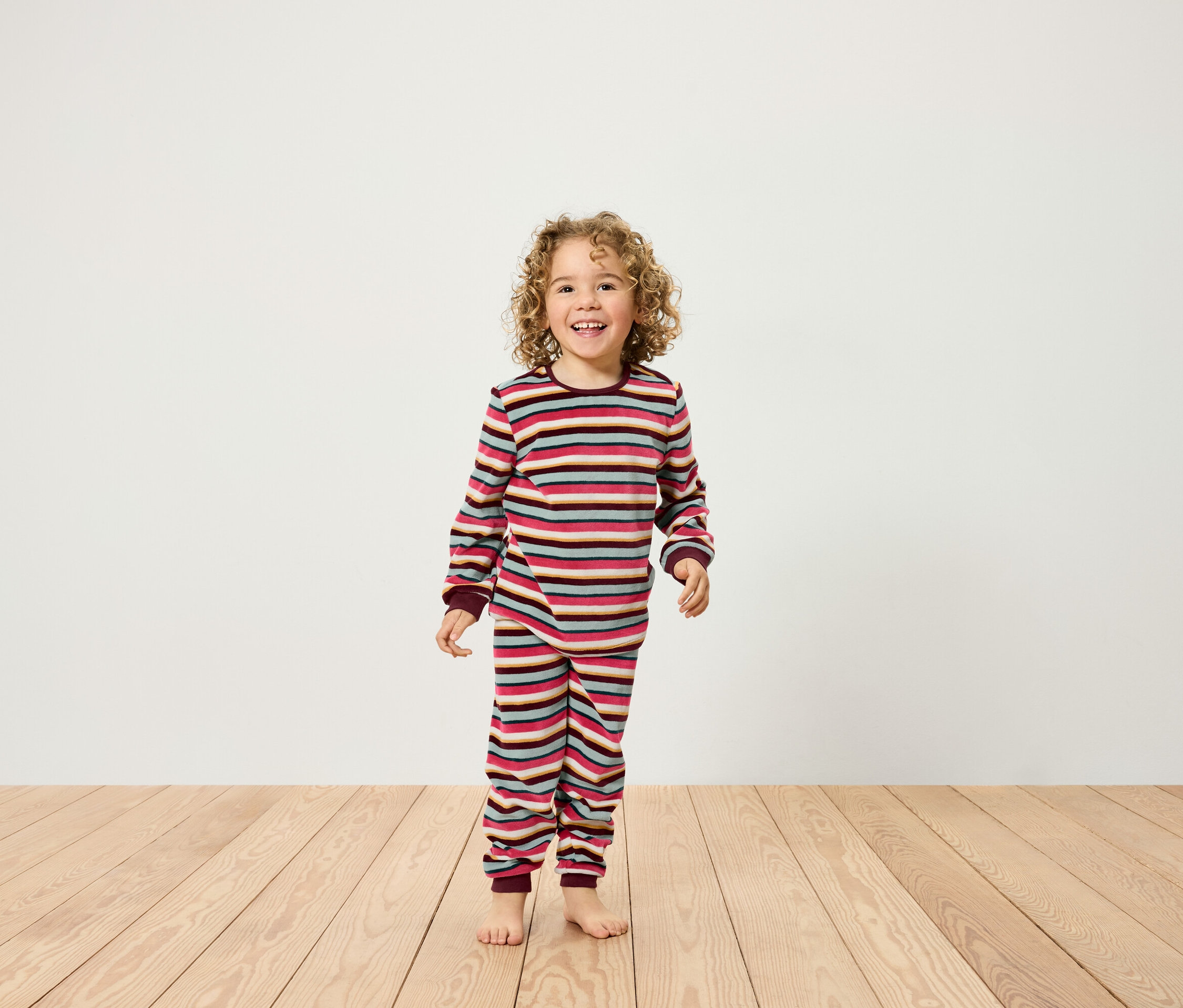 Usmívající se dítě s kudrnatými vlasy stojí na dřevěné podlaze a má na sobě pruhované pyžamo.