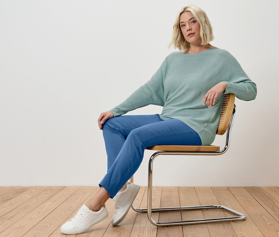 Blondýna sedí na židli a má na sobě modré strečové kalhoty a šalvějový svetr z jemného úpletu.