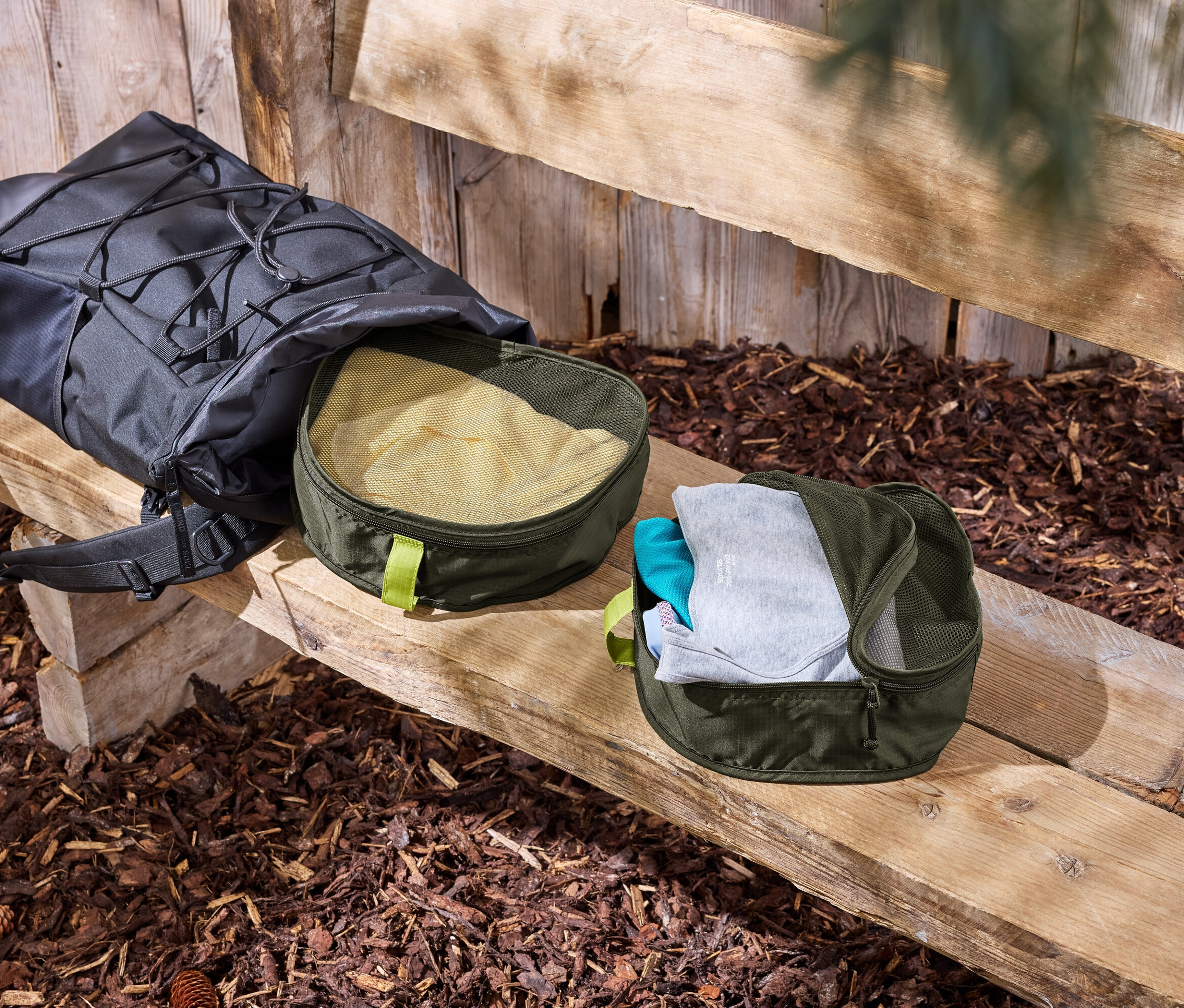 Černý batoh leží na dřevěné lavici vedle dvou zelených organizérů na oblečení.