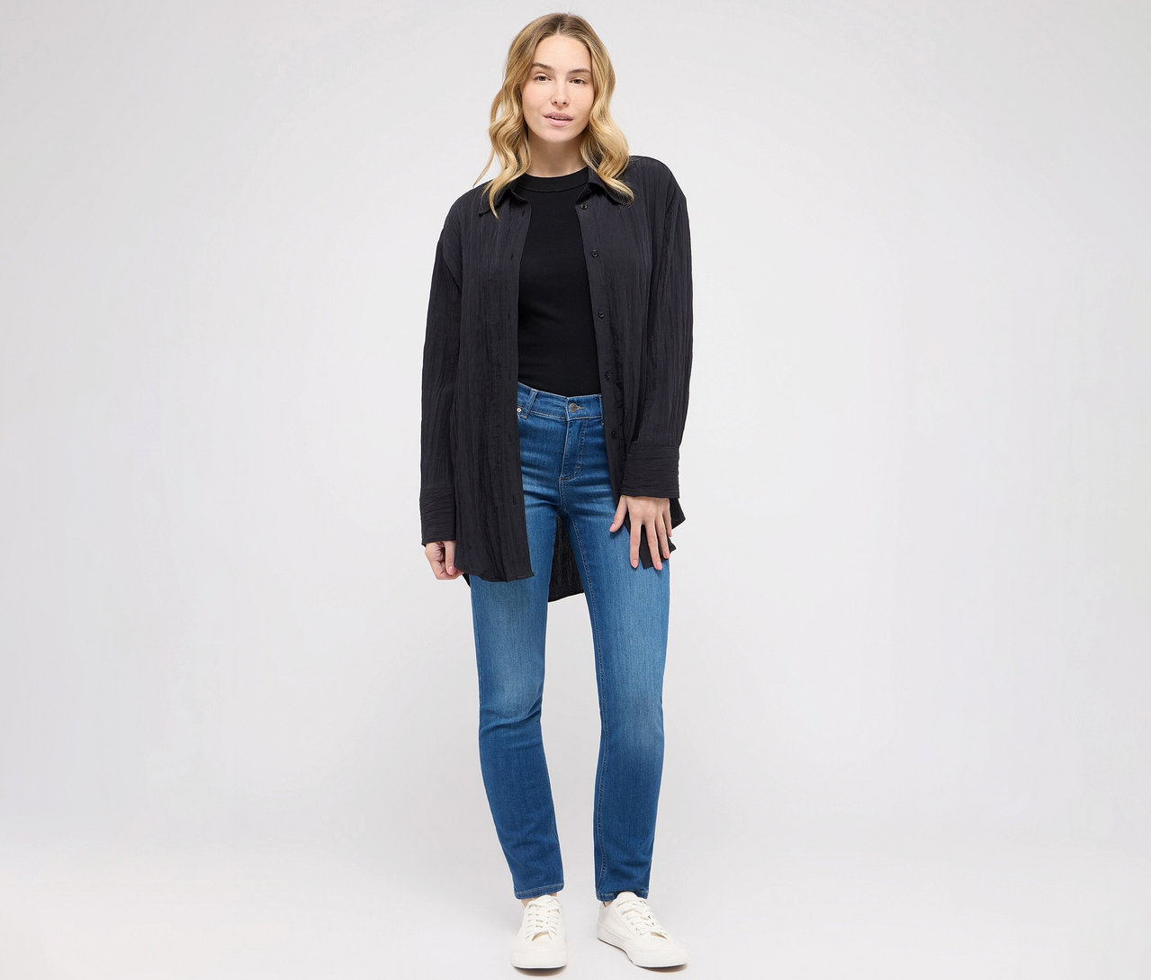 Žena pózuje ve slim fit džínách Angels »Cici« středně modré barvy, černém tričku a halence.