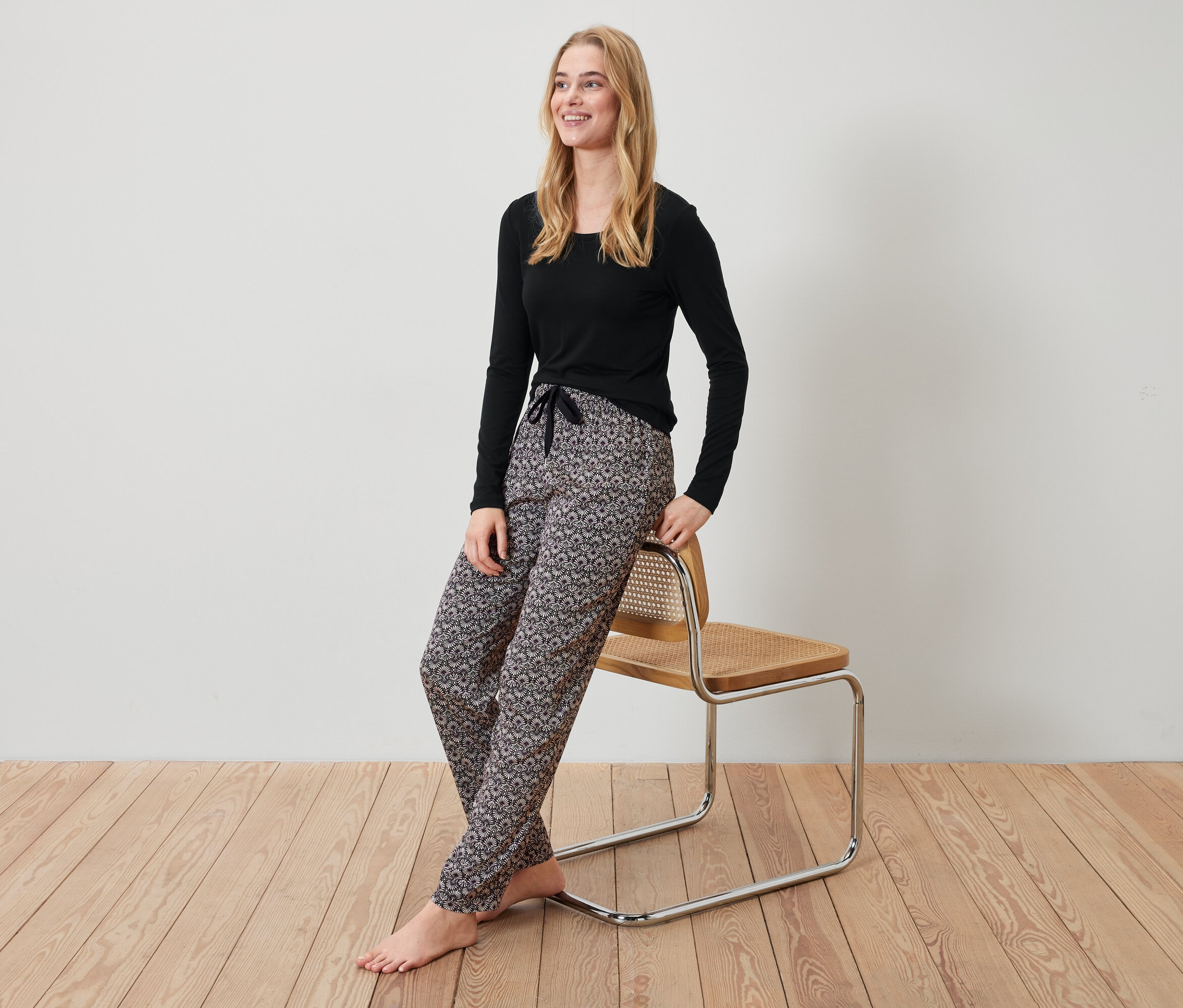Blondýna v černém tričku a vzorovaných kalhotách opřená o židli.