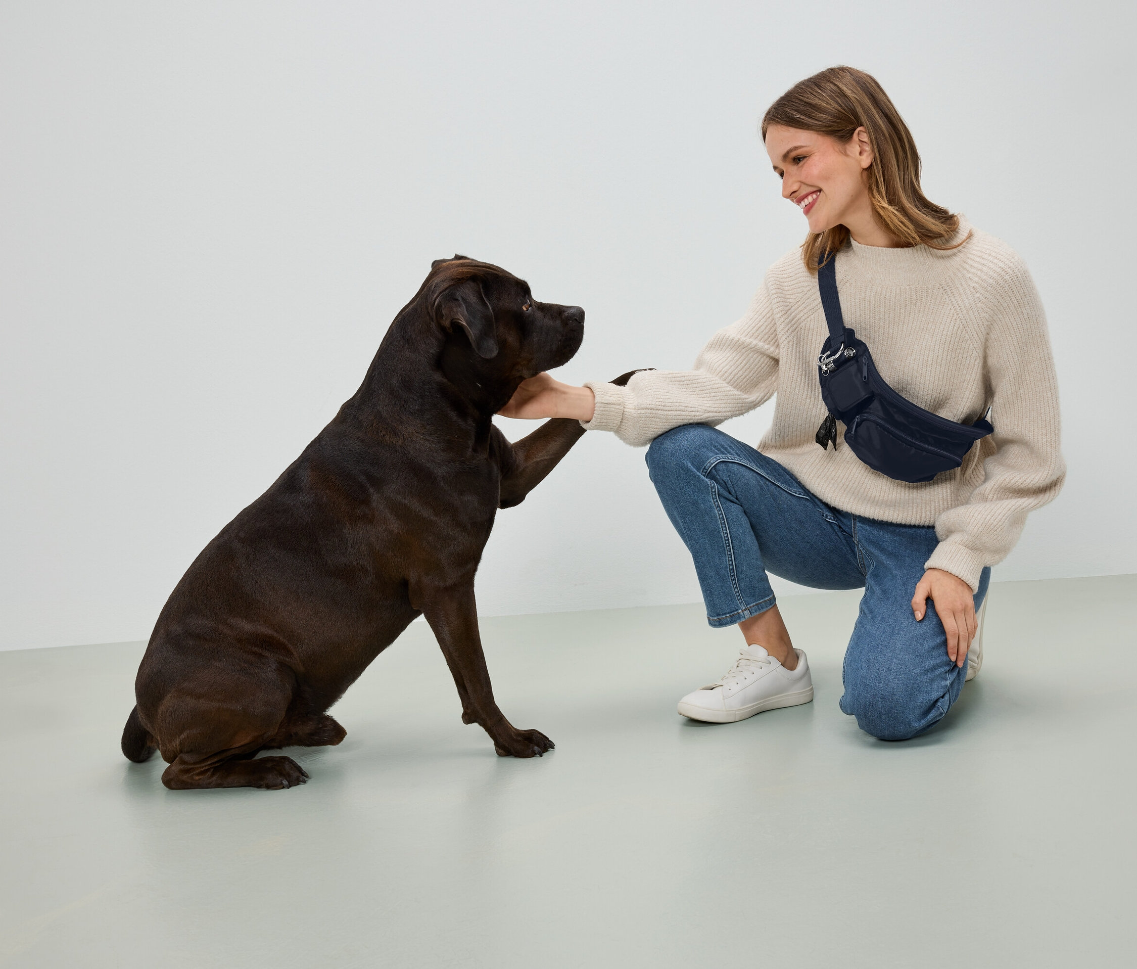Žena v béžovém svetru a modrých džínách klečí a drží tlapu tmavě hnědého psa. Má na sobě tmavě modrou ledvinku.