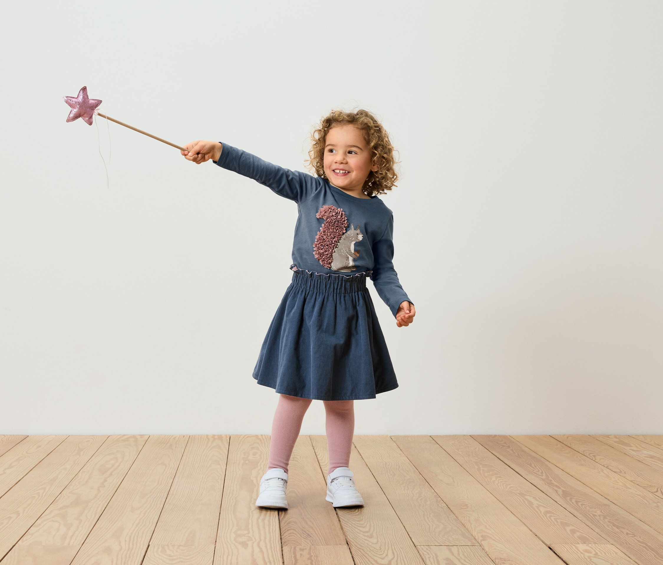 Dívka s kudrnatými vlasy v modrých šatech drží kouzelnou hůlku s hvězdou.
