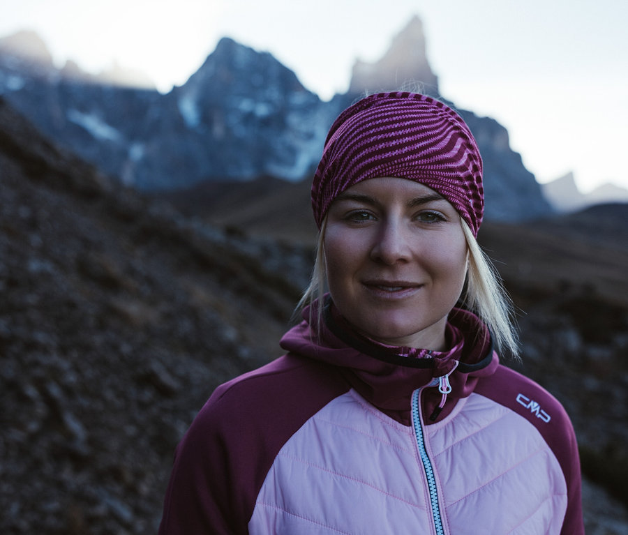 Blondýna v hybridní bundě CMP beere a pruhovaném šátku na hlavě stojí v horách.