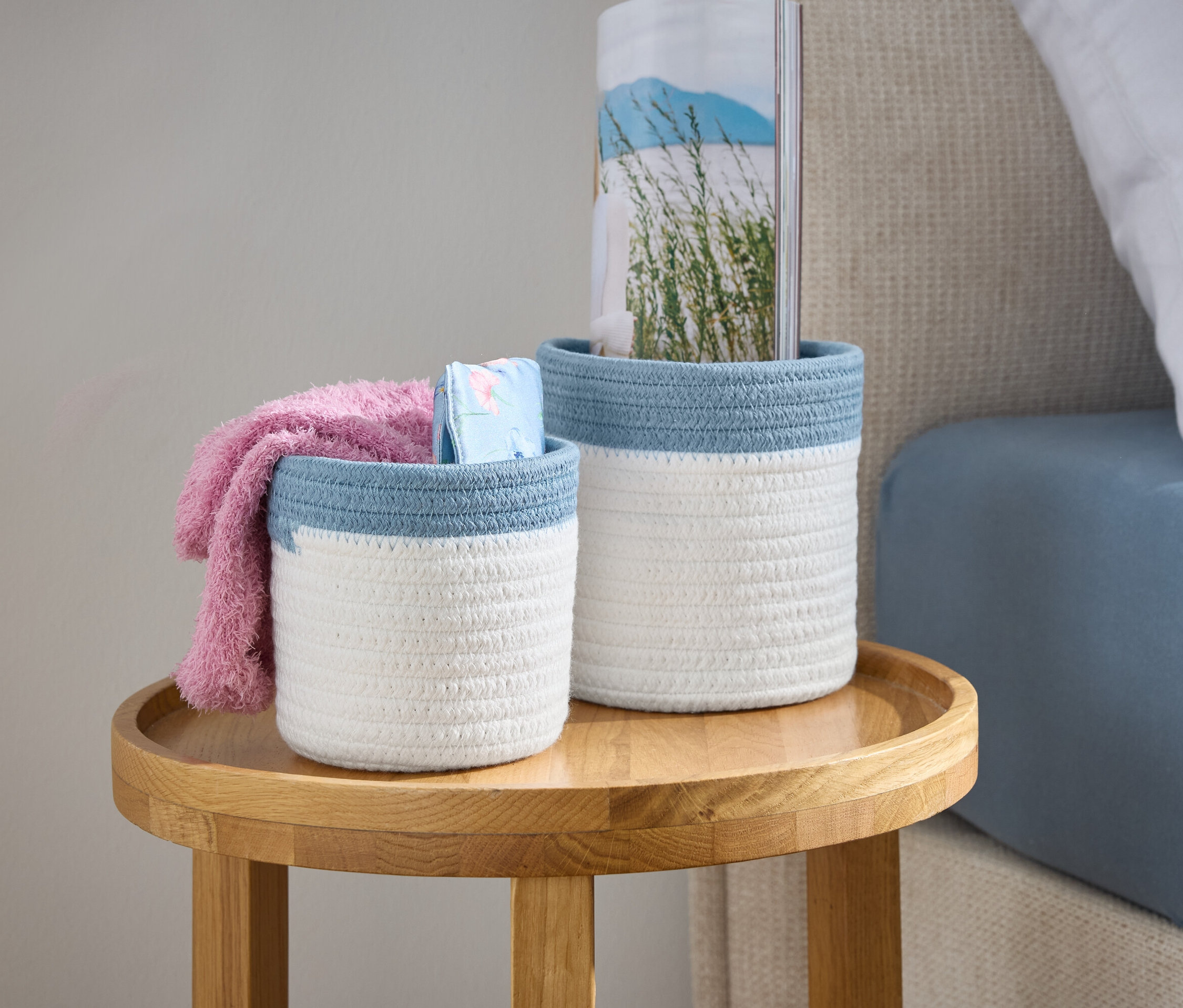 Dva bílé pletené košíky s modrým lemem stojí na dřevěném stolku vedle postele.