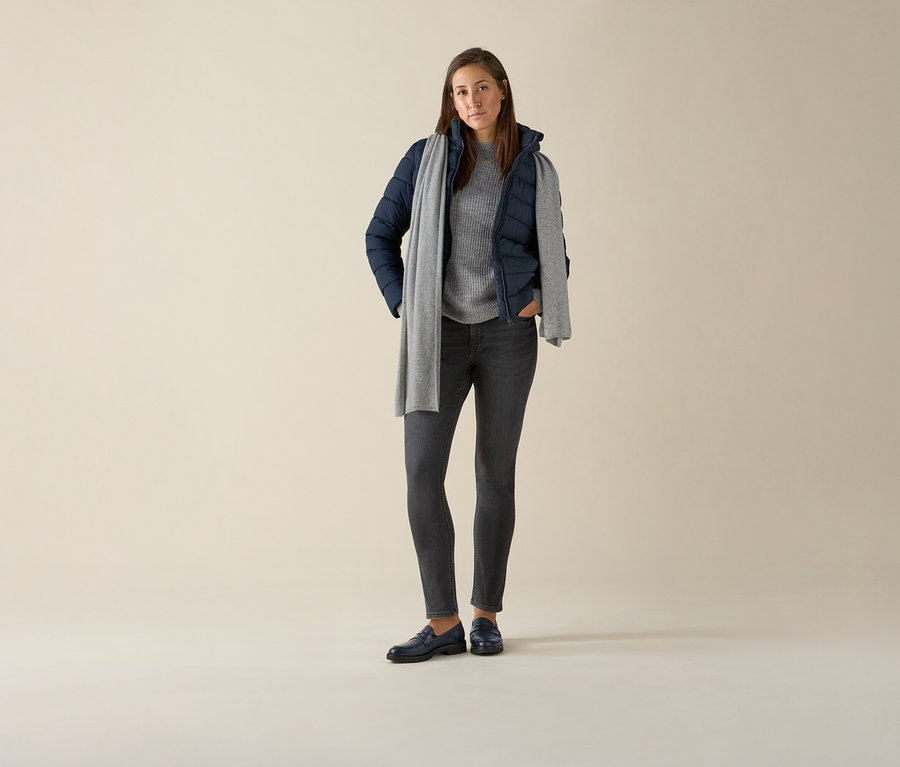 Žena stojící a má na sobě džíny slim – střih »Emma«, grey denim, šedý svetr, tmavě modrou bundu a šedou šálu.