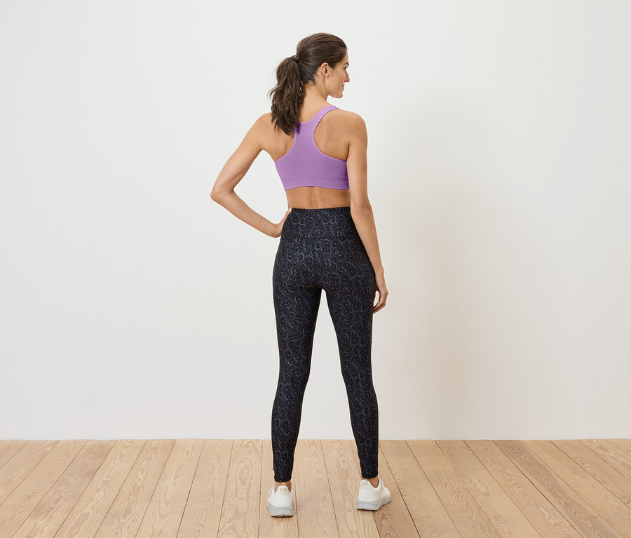 Žena stojí zády, má na sobě fialovou sportovní podprsenku a černé legíny.