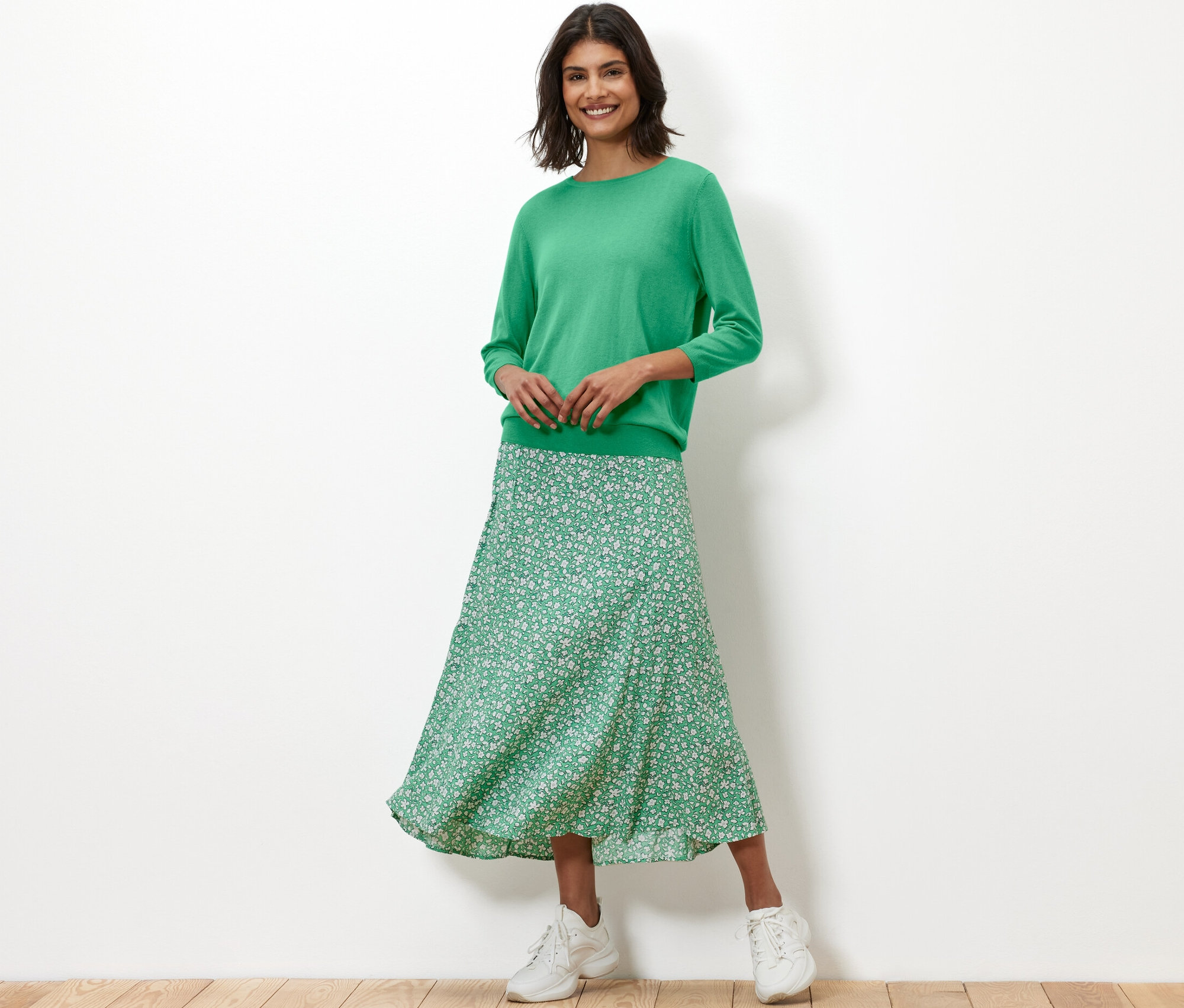 Žena má na sobě zelený svetr z jemného úpletu s hedvábím a květinovou sukni.