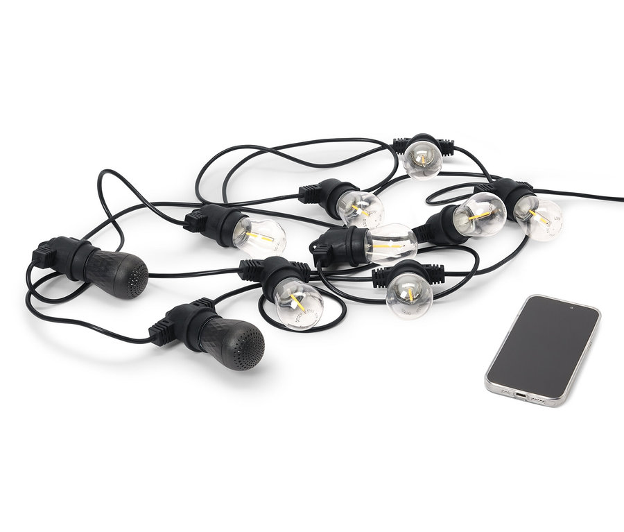 Černý venkovní světelný řetěz s LED a 2 reproduktory Bluetooth® leží vedle mobilního telefonu na bílém pozadí.