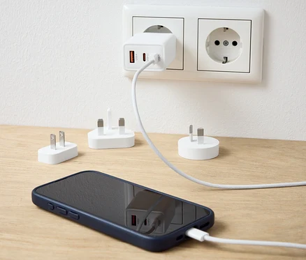 Bílý USB cestovní adaptér zapojený do zásuvky nabíjí mobilní telefon. Na stole jsou další adaptéry.